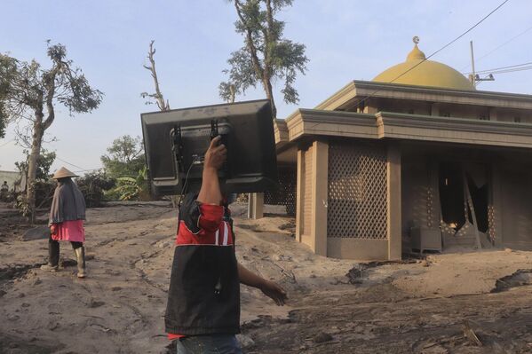 مردی در حالی که از کنار مسجدی که تا حدی در خاکستر آتشفشانی ناشی از فوران کوه سمرو در روستای کاجار کونینگ در لوماجانگ، جاوه شرقی، اندونزی مدفون شده است و تلویزیون را حمل می‌کند، - اسپوتنیک افغانستان  