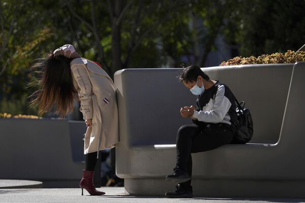زنی که برای جلوگیری از شیوع ویروس کرونا ماسک صورت می‌بندد، بدن خود را در کنار زنی نقابدار که روی نیمکتی بیرون یک مرکز خرید در پکن نشسته است، به سمت عقب کش می دهد. - اسپوتنیک افغانستان  