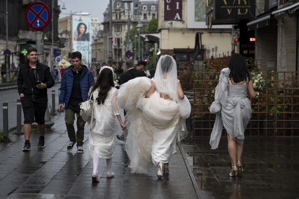 عروس در حالی که بعد از بارندگی در مرکز شهر بخارست، رومانیا، شنبه، 27 می 2023 راه می رود، لباس خود را حمل می کند - اسپوتنیک افغانستان  