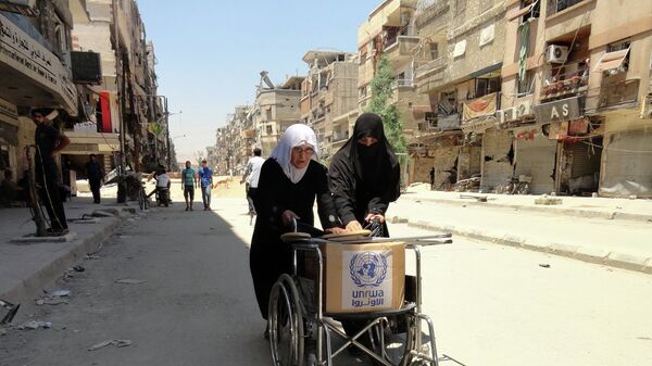 Residents of Syria's Yarmuk Palestinian refugee camp, south of Damascus, use a wheelchair to carry a box of goods distributed by the United Nations Relief and Works Agency (UNRWA) on July 17, 2014 - اسپوتنیک افغانستان  